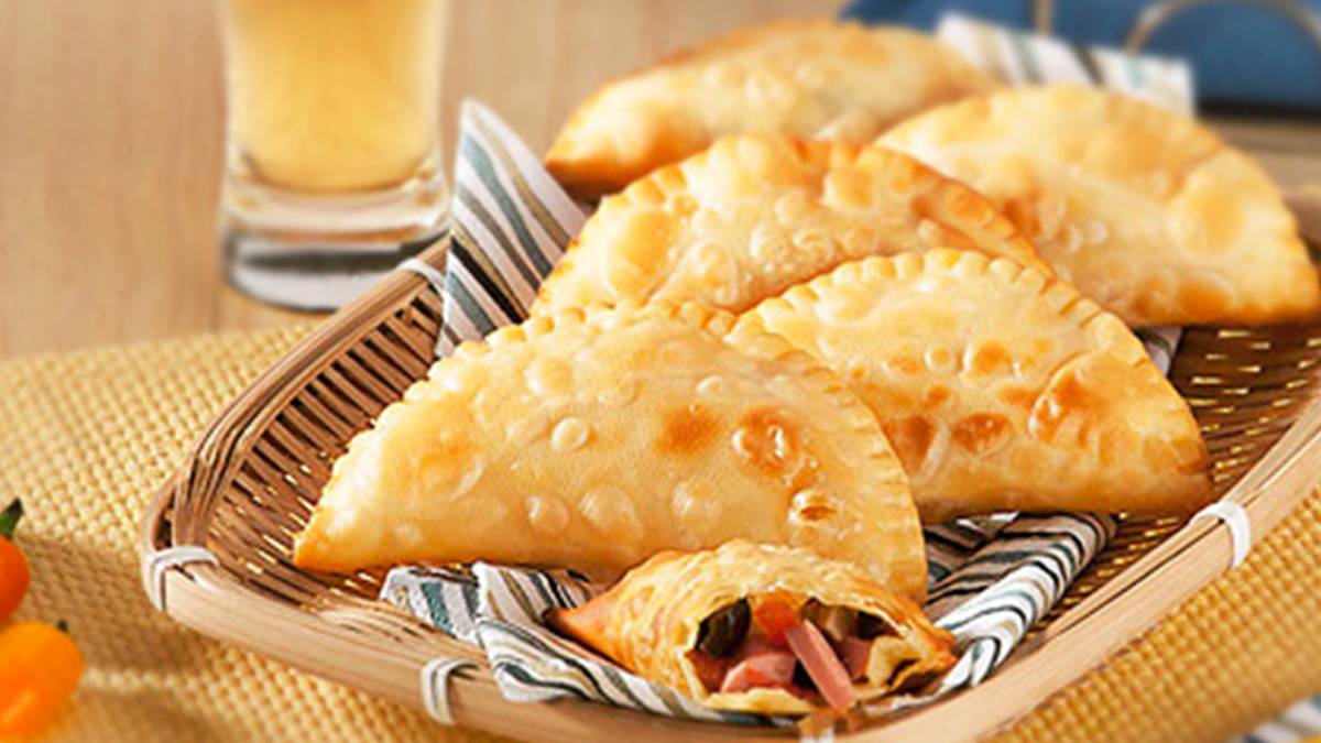 Pastéis de salsicha de frango em uma cestinha retangular forrada com um guardanapo listrado azul e cinza. Ao fundo da foto, um copo de suco meio amarelado, aparentado ser suco de abacaxi.