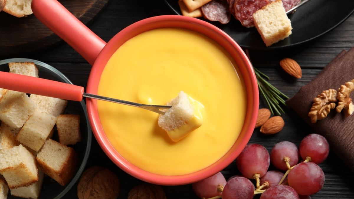 Imagem destaca a receita de fondue de queijo servido em uma panela vermelha. Ao lado, os acompanhamentos para fondue: pães torradinhos, uvas e salame fatiado.
