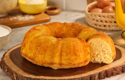 Bolo de milho servido em uma tábua de madeira rústica parecida com um tronco de árvore. O bolo foi cortado e há uma fatia na direita mostrando o interior do bolo, que é meio amarelado e visualmente fofinho. Ao fundo da imagem, do lado esquerdo, uma garrafa de café antiga esmaltada de cor amarela e uma xícara esmaltada também amarela. Mais atrás, uma cesta com ovos caipiras. Do lado esquerdo, uma tábua de madeira com uma tigela amarela redonda com coco ralado e outra maior com farinha de trigo. Mais à frente, do lado esquerdo do bolo, uma tigela com açúcar refinado.
