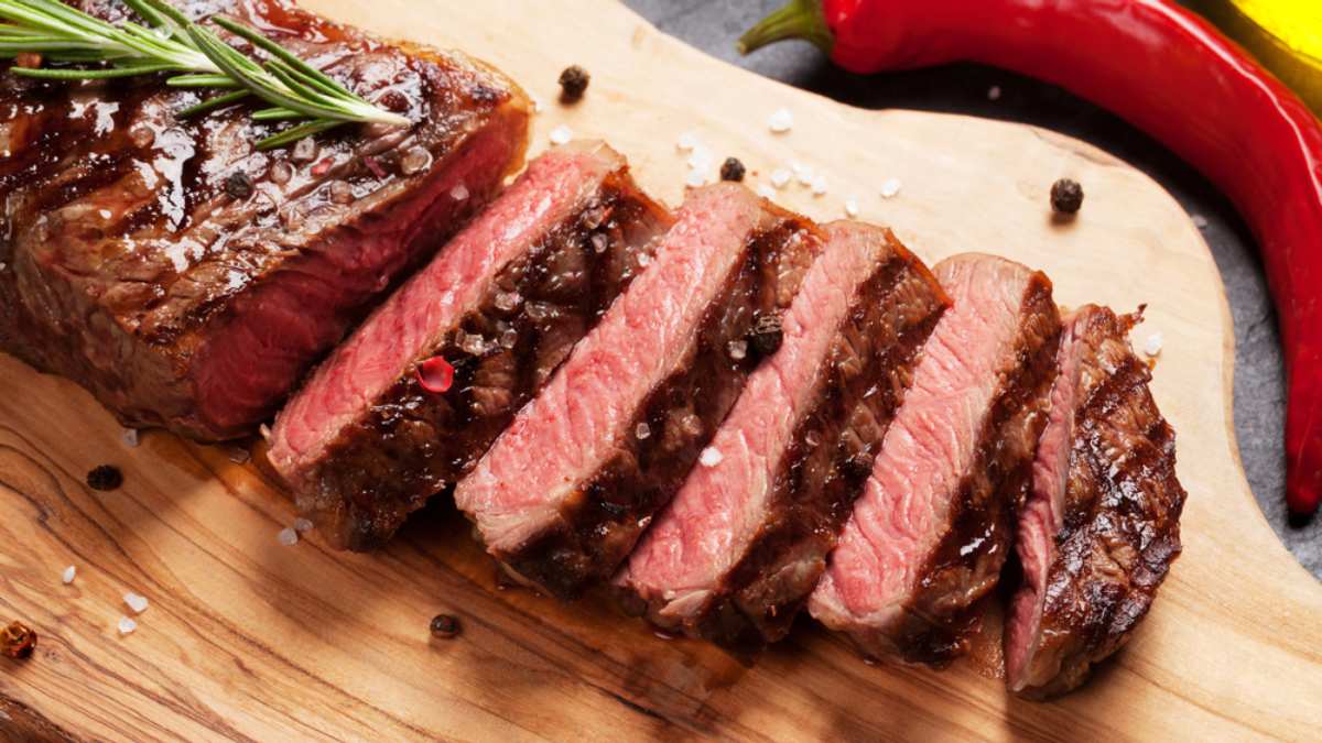 Imagem destaca uma carne de churrasco mal passada e fatiada, com uma crosta dourada por fora, servida em tábua de madeira.