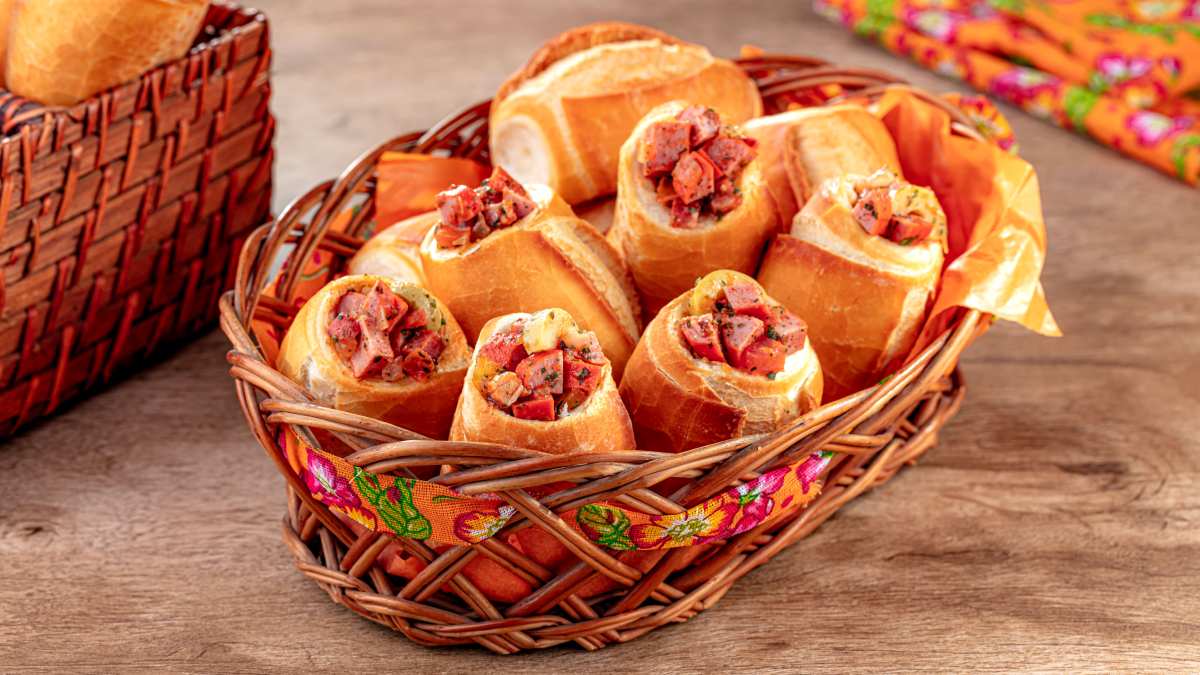A imagem destaca uma cesta decorada com tecido de estasmpa junina com várias unidades de buraco quente de salsicha dentro.