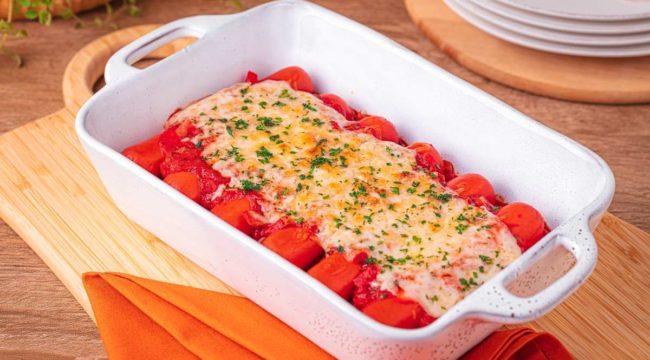 Na imagem, vemos uma travessa retangular com salsichas à parmegiana recém-saídas do forno. Cada salsicha está coberta com molho de tomate encorpado e queijo mussarela derretido, formando uma camada dourada e apetitosa sobre cada uma. O prato está posicionado sobre uma superfície de madeira escura, destacando as cores vibrantes do molho vermelho e do queijo derretido. A cena transmite uma sensação de comida caseira e reconfortante, pronta para ser servida em um jantar em família.